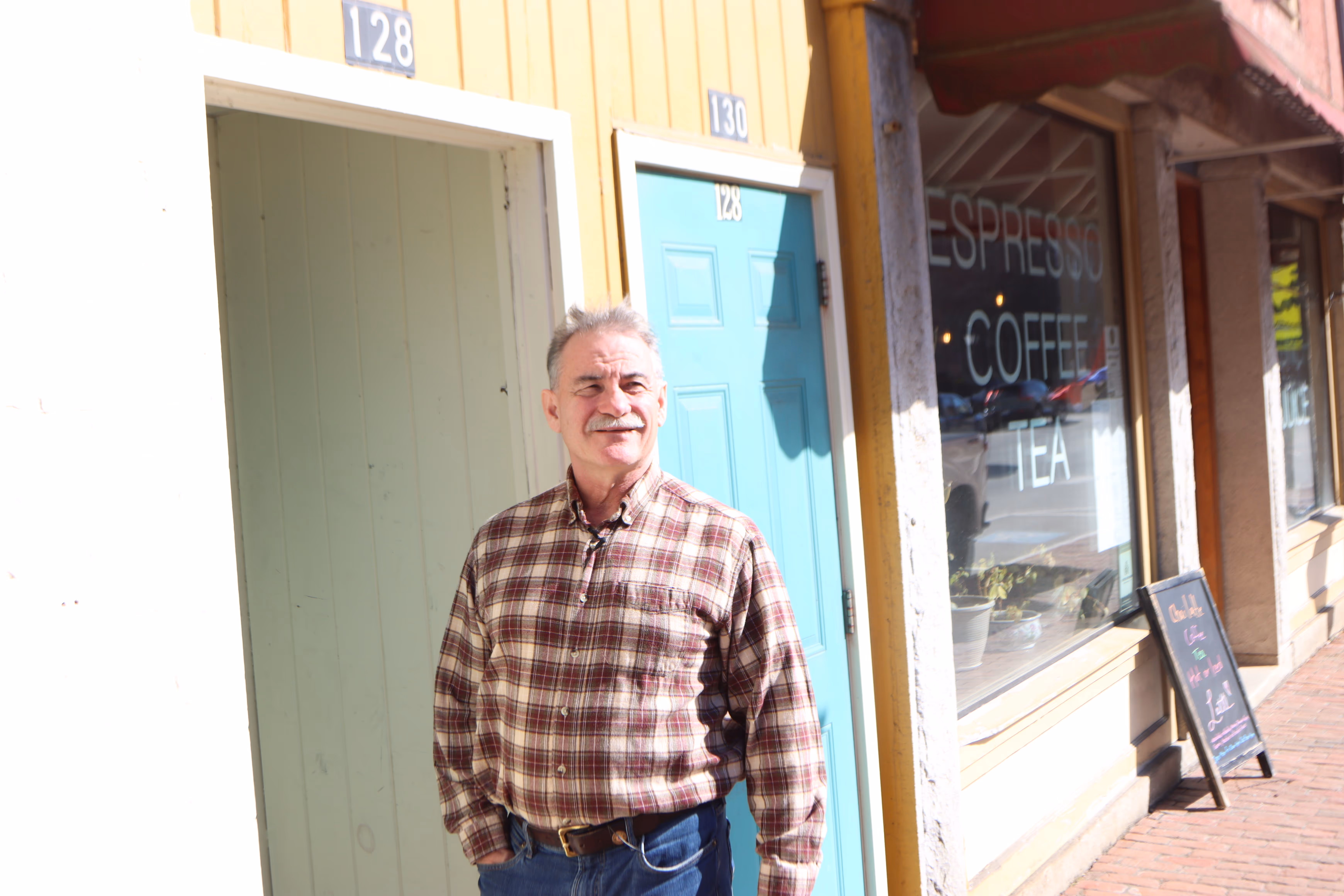 Smiling man in a plaid shirt standing outside a yellow building.