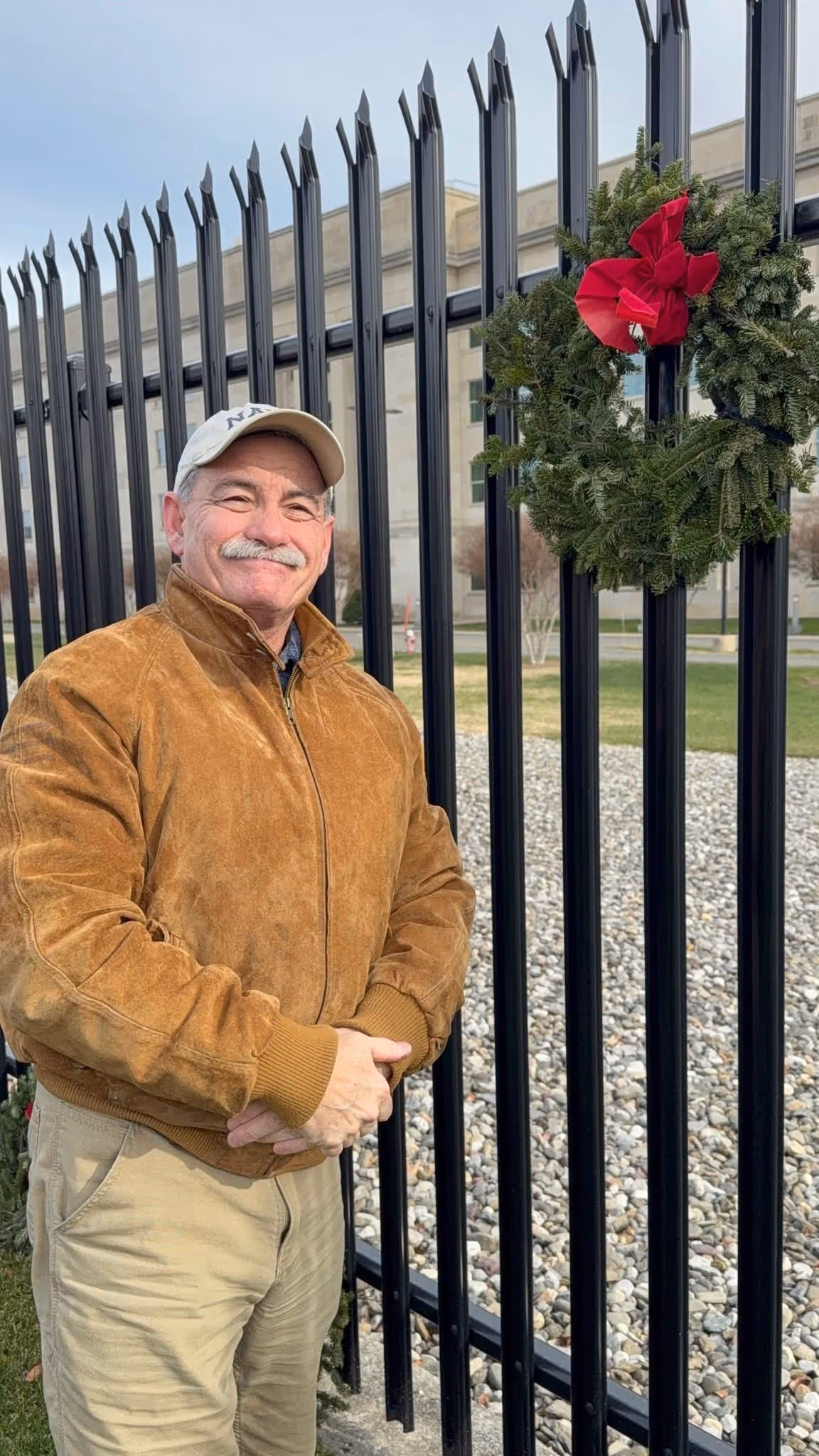 Smiling man wearing a brown jacket and beige cap standing by a black iron fence.