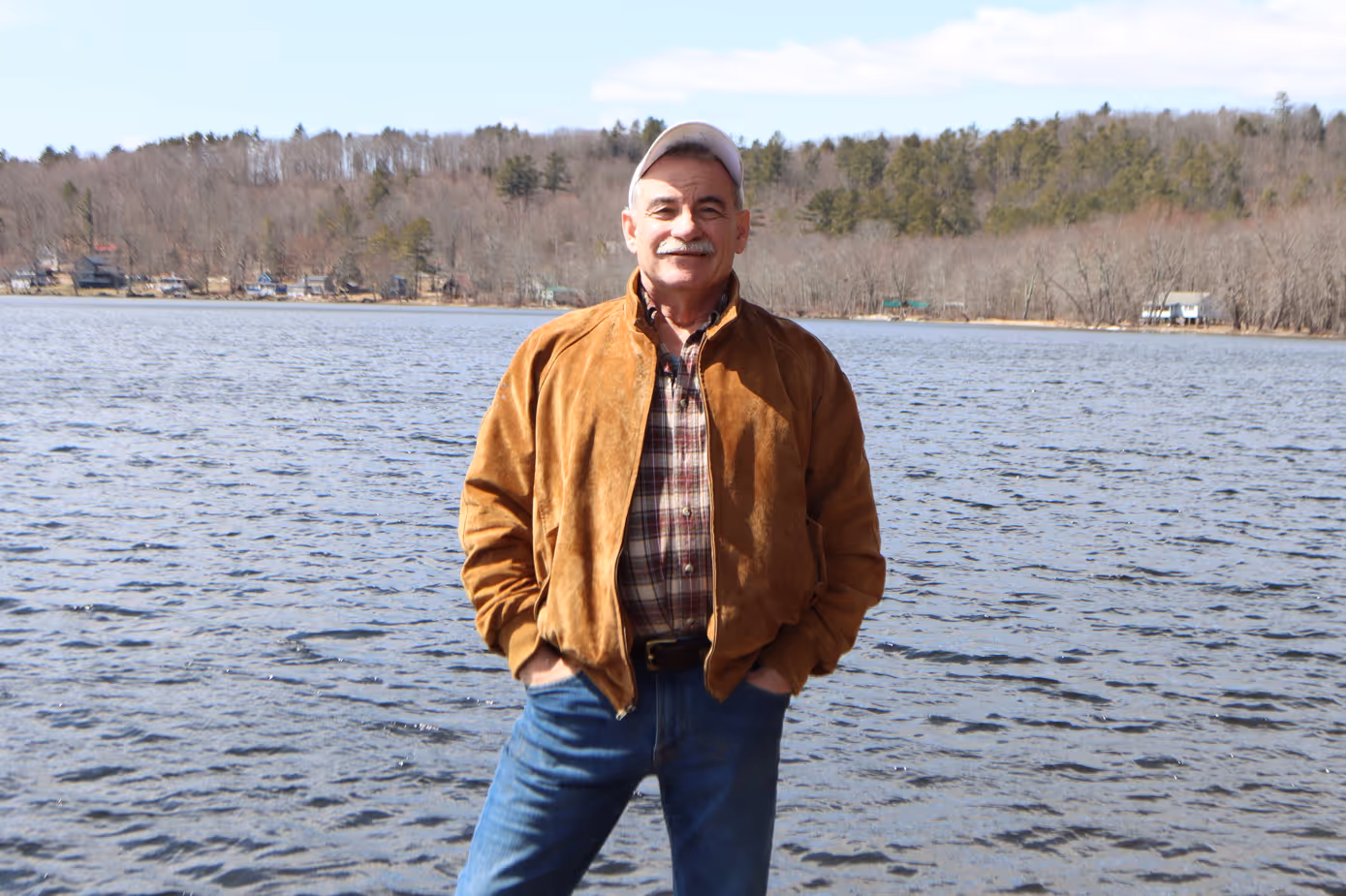 Smiling middle-aged man in brown jacket and cap standing by a lake.