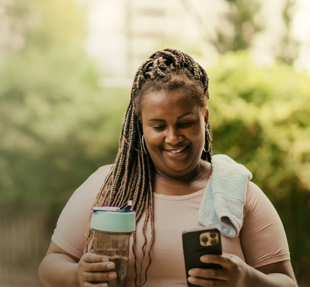 Woman holding a water bottle and phone, smiling while outdoors with a towel draped over her