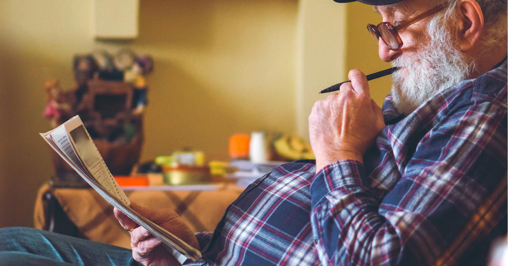 An elderly man in glasses reads a newspaper, holding a pen thoughtfully, seated indoors.