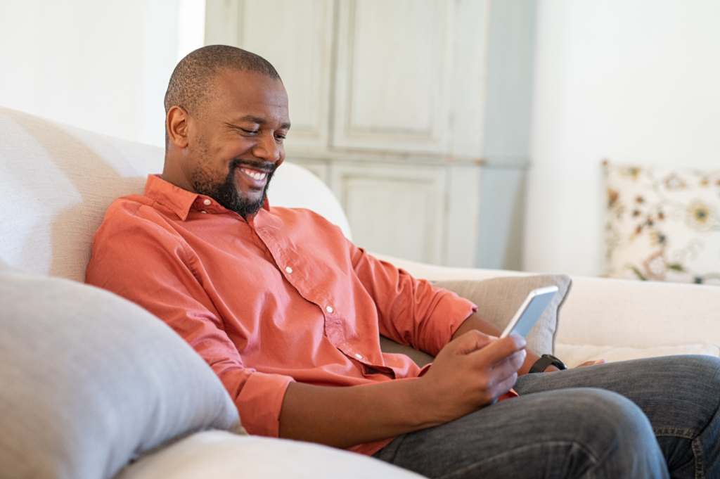 Man smiling while looking at his smartphone, sitting on a couch in a bright room.
