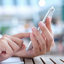 A hand holding a smartphone, tapping the screen, on a wooden table in an outdoor setting.