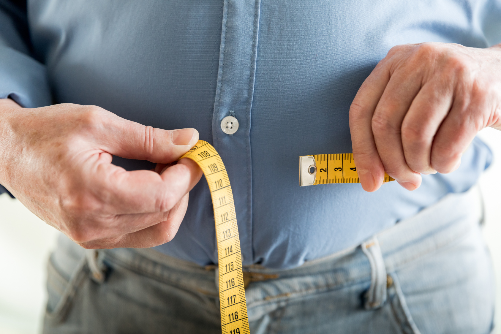 A person measures their waist with a tape measure while wearing a button-up shirt.