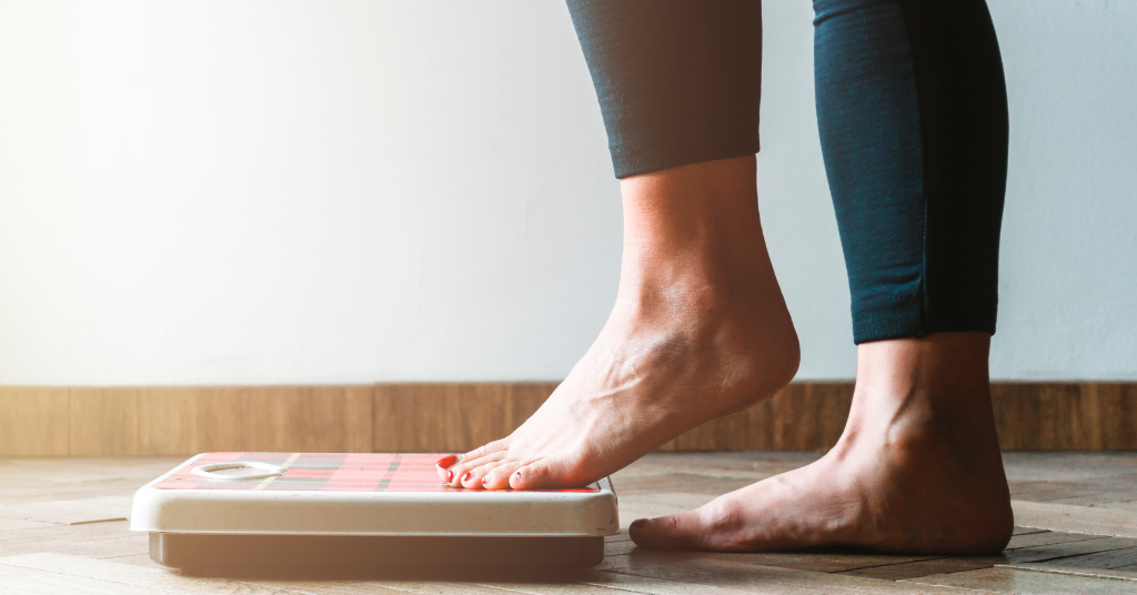 A person in leggings weighs themselves on a bathroom scale with one foot.