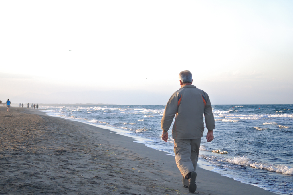 An older man walks along a sandy beach, with gentle waves and a soft sunset in the background.