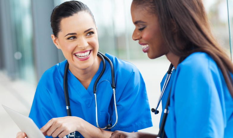Two smiling healthcare professionals in blue scrubs interact while using a tablet.