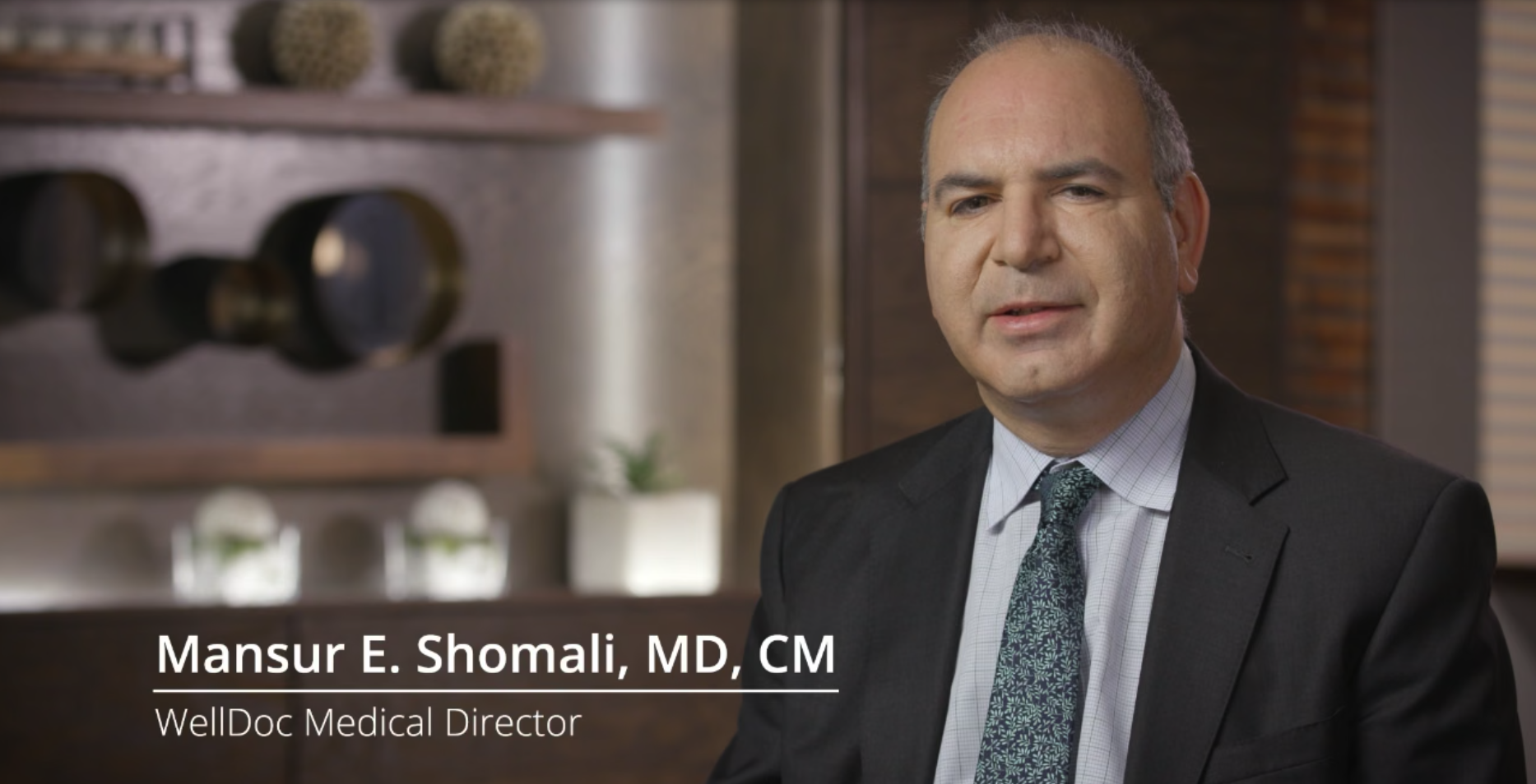A man in a suit speaking in a well-lit room with modern decor, identified as a medical director.