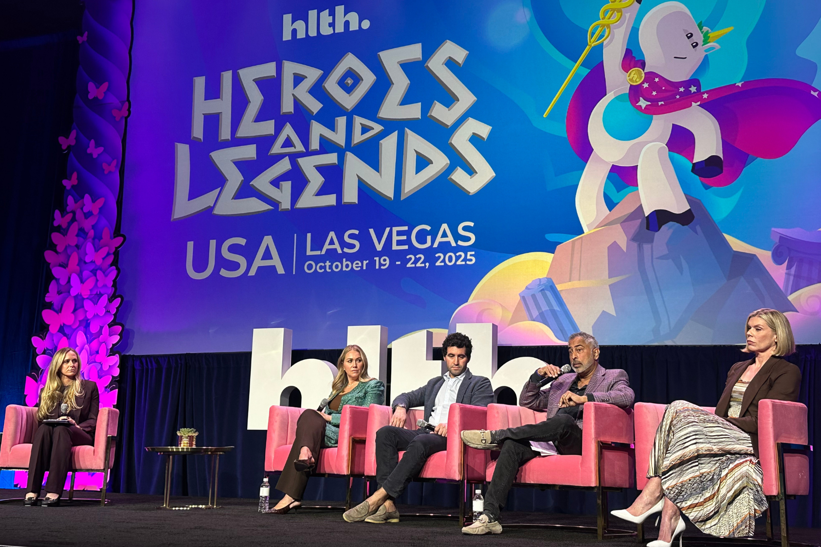 Panel discussion at HLTH 2025 with four speakers on stage, colorful backdrop displaying event theme.