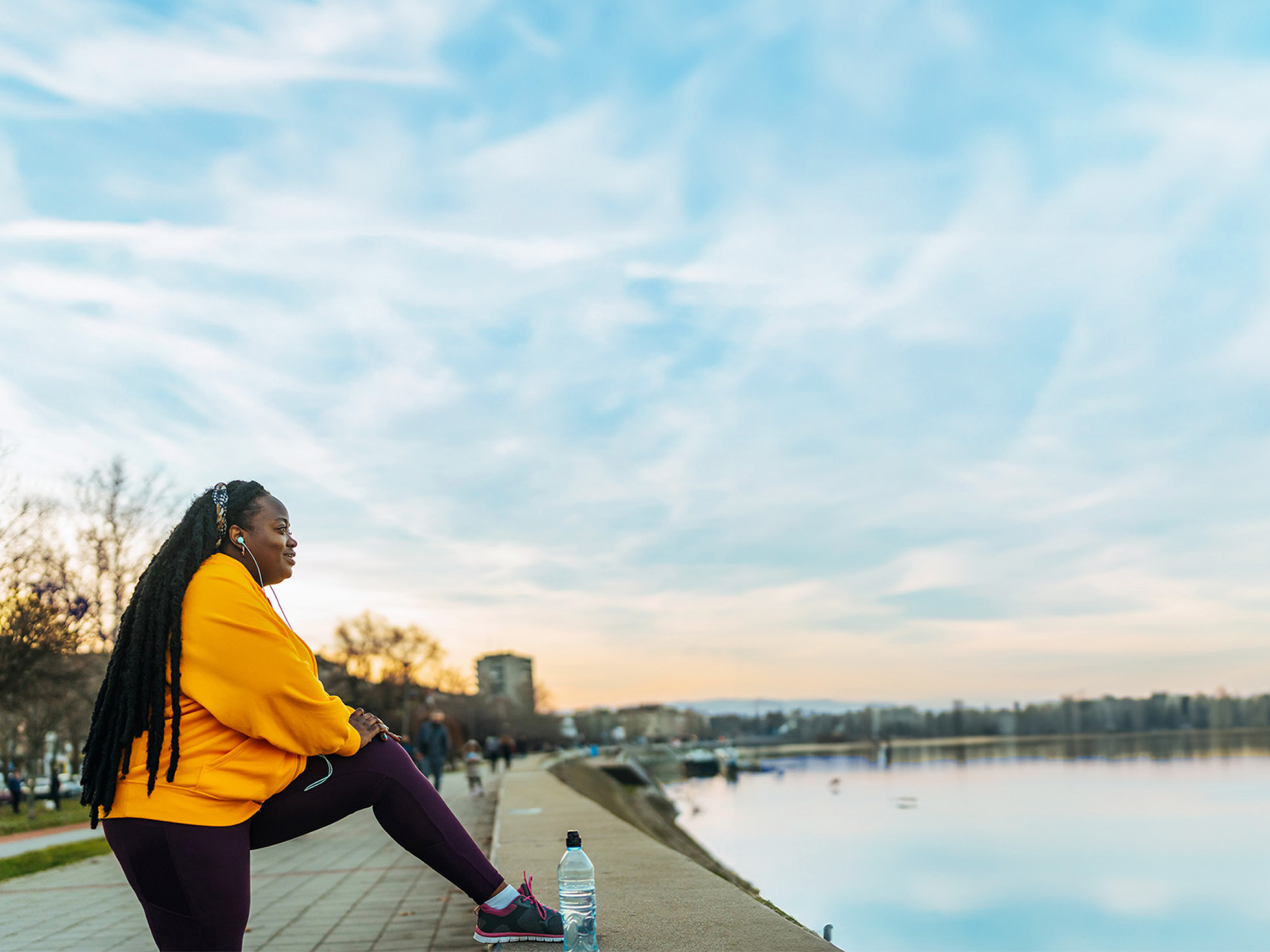 A woman in a yellow hoodie sits by a lake, stretching, with a water bottle nearby.