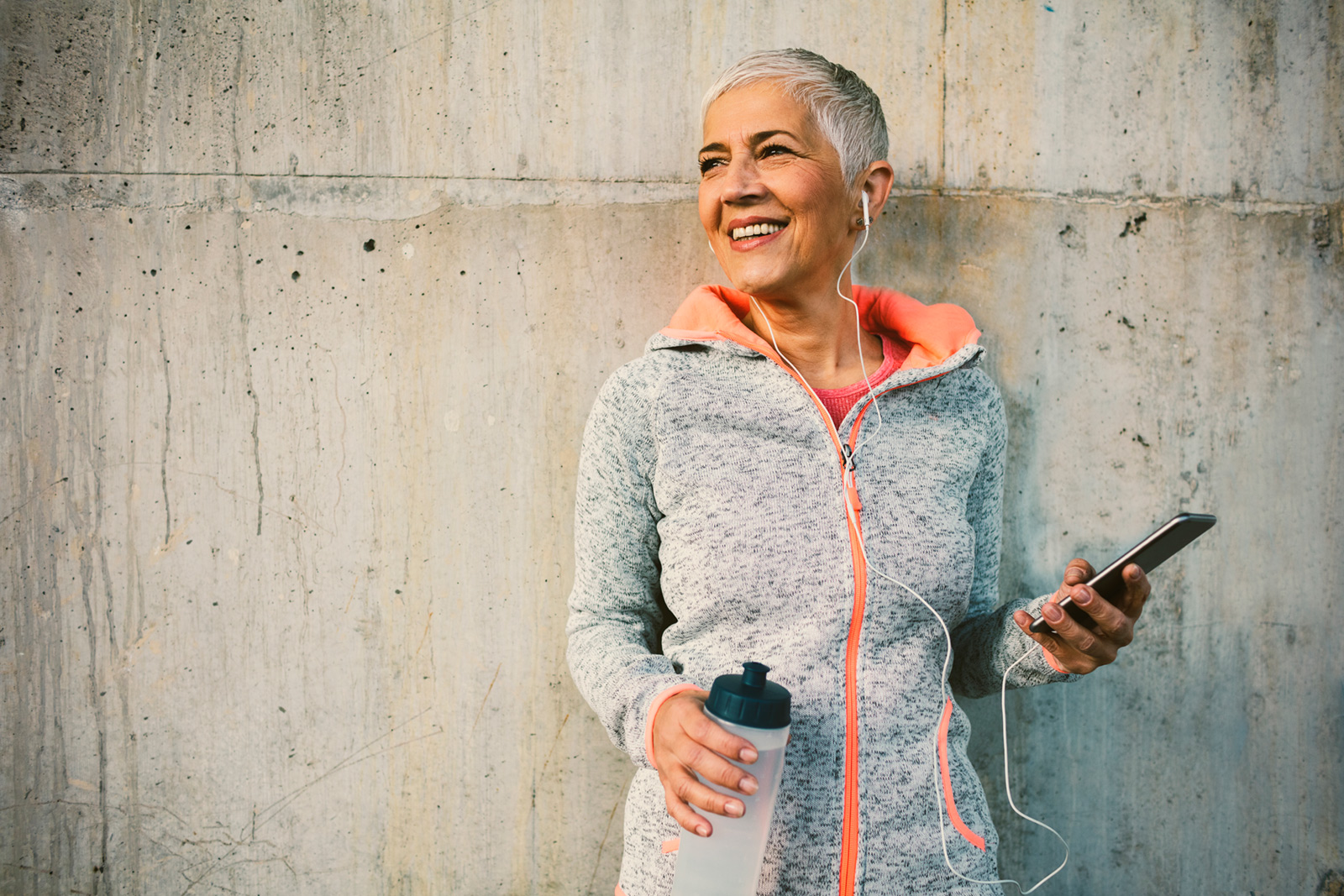 Smiling older woman in athletic wear holds a phone and water bottle against a concrete wall.