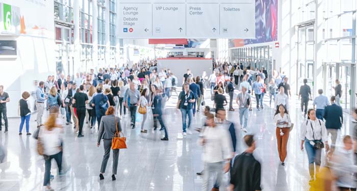 A busy convention center with people walking and various directional signs above.
