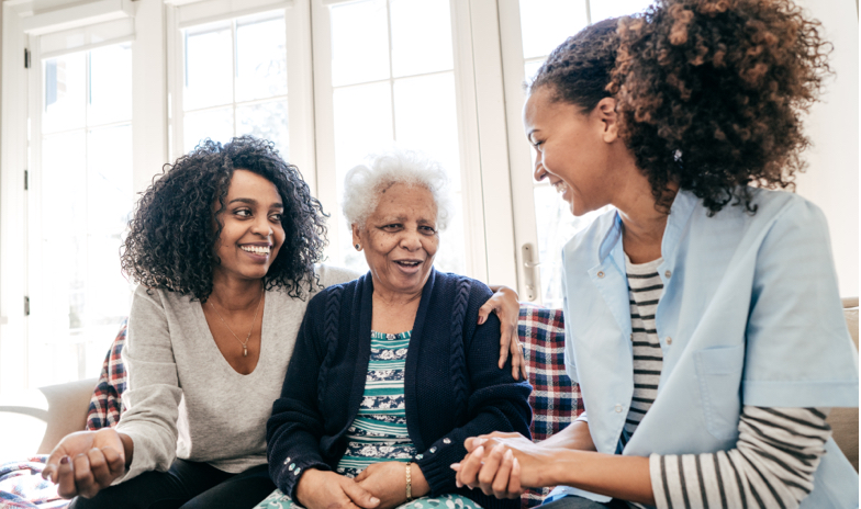 Three women of different ages share a moment, smiling and engaged, in a cozy, well-lit room.