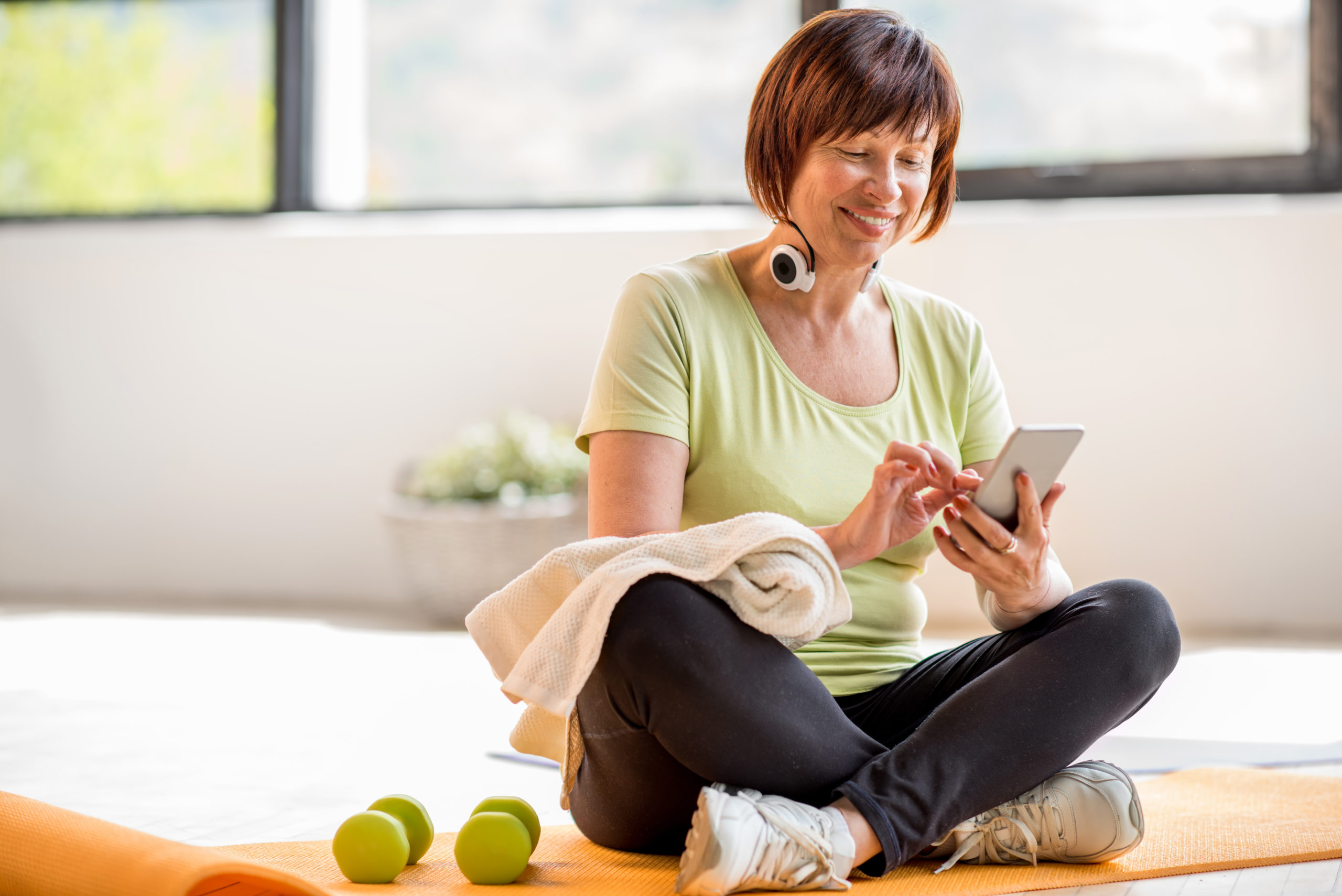 A woman sitting on a mat, smiling at her phone, with dumbbells nearby and a towel in hand.