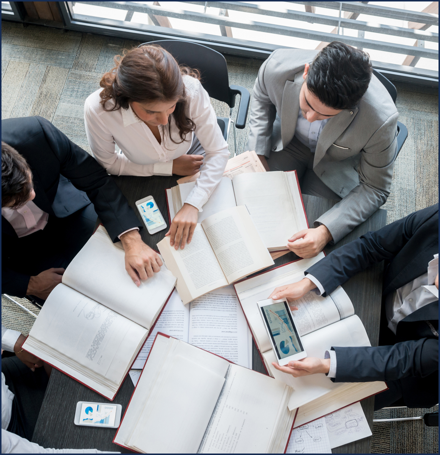 A group of professionals collaborates over open books and devices in a modern office setting.