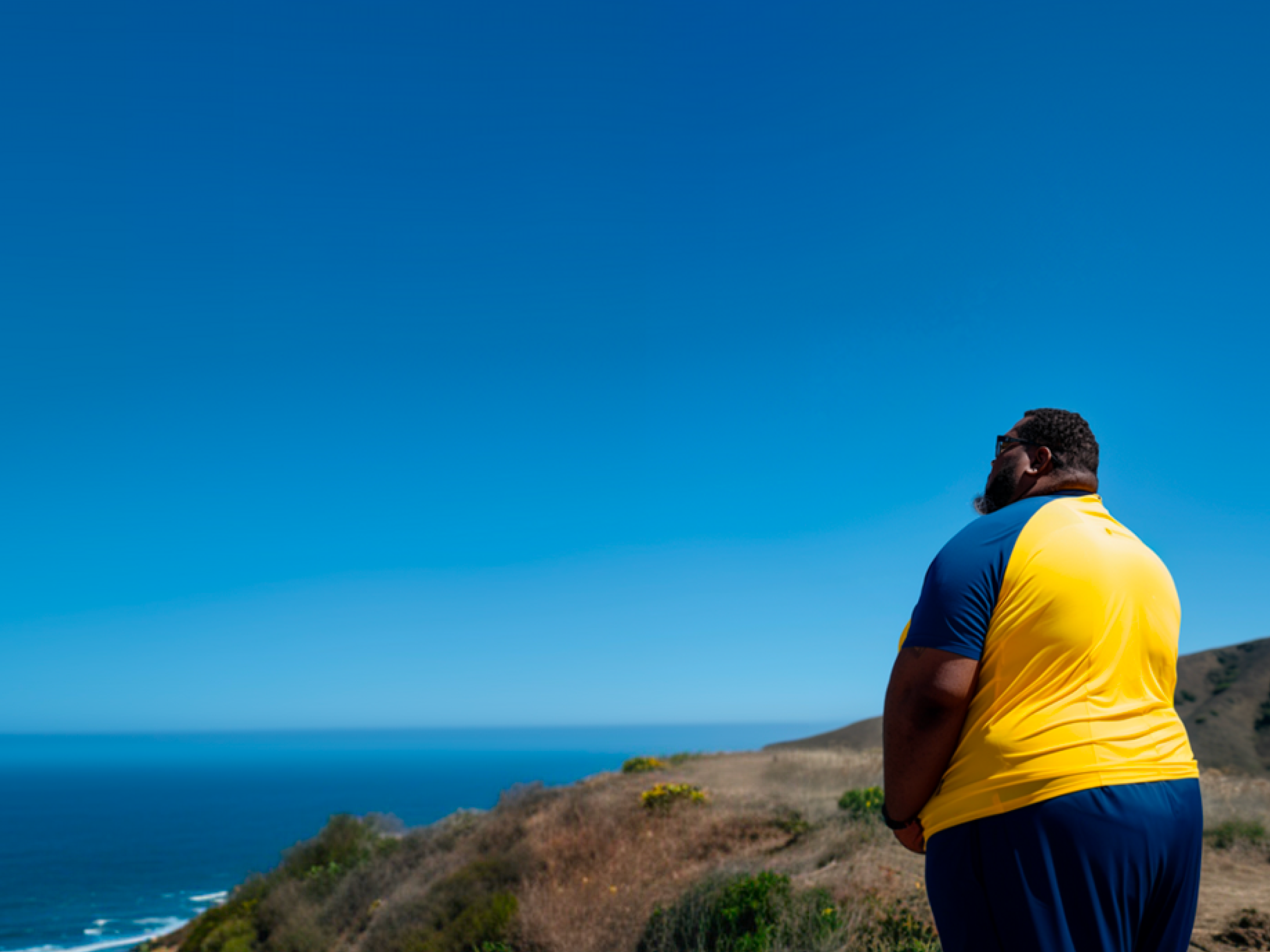 A person stands overlooking the ocean, wearing a yellow and blue sports shirt, on a hillside.