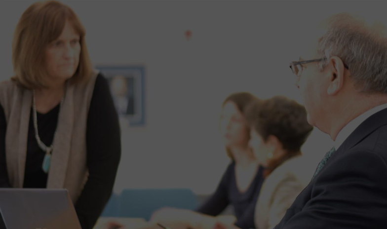 A group of professionals in a meeting, with a focus on a woman speaking to a man.