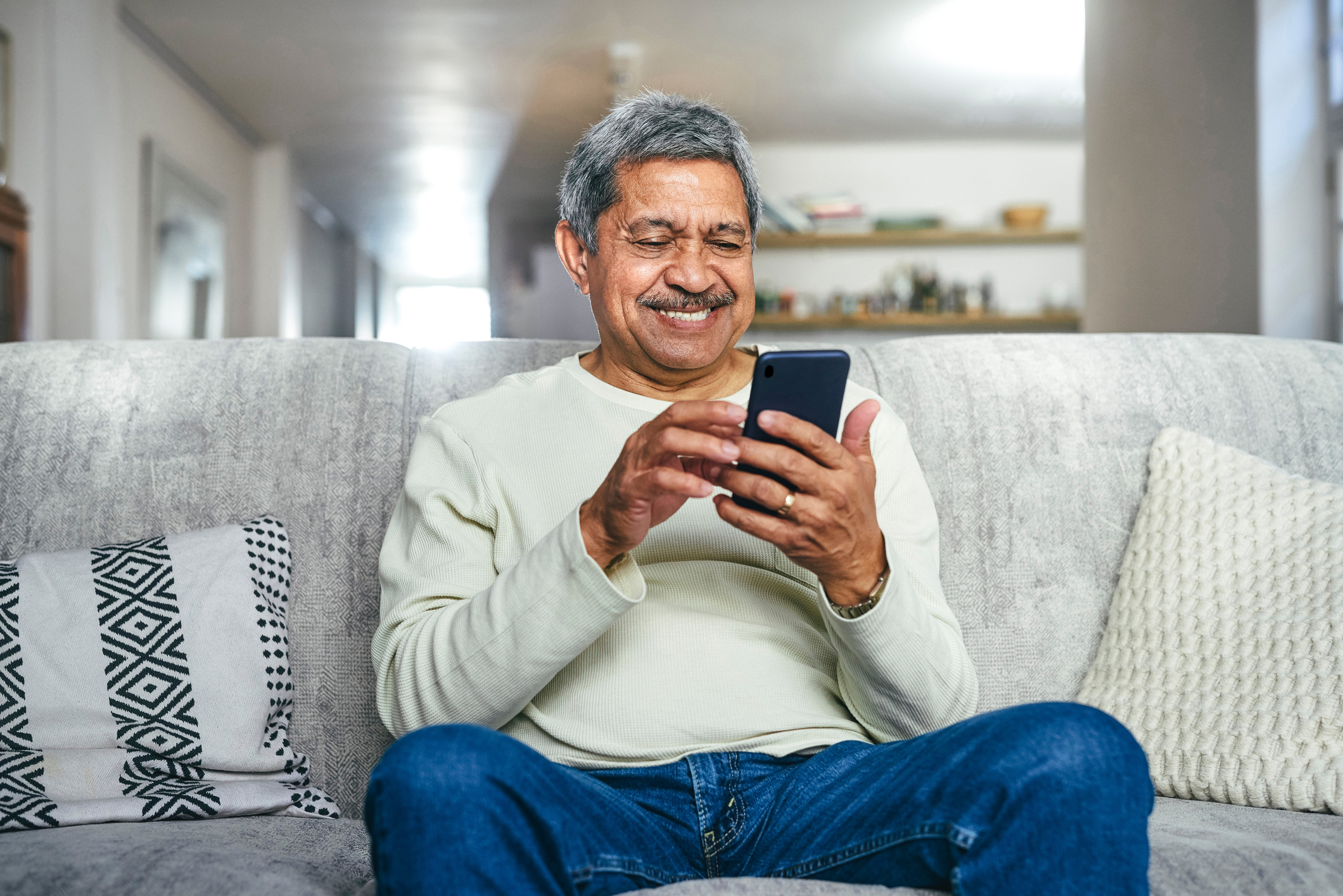Older man sitting on a couch, smiling while texting on his smartphone. Soft, cozy environment.
