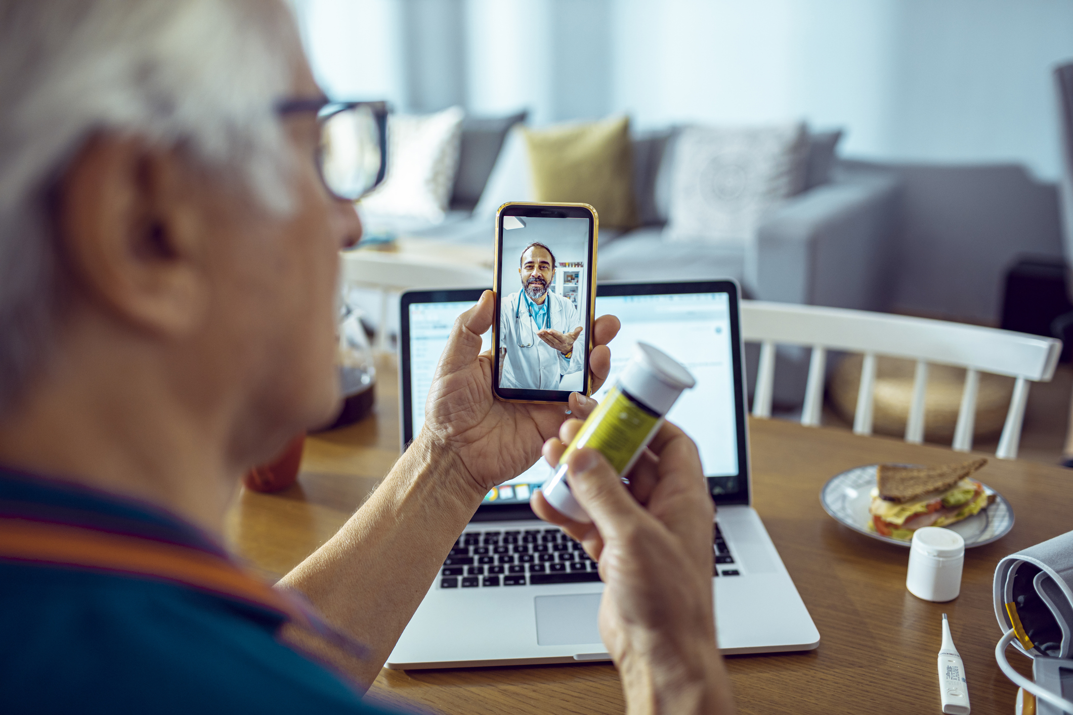 An older man on a video call holds a pill bottle, with a laptop and food on the table.
