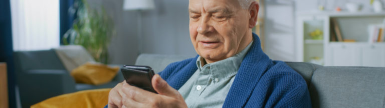 An older man in a blue sweater uses a smartphone while sitting on a gray couch.