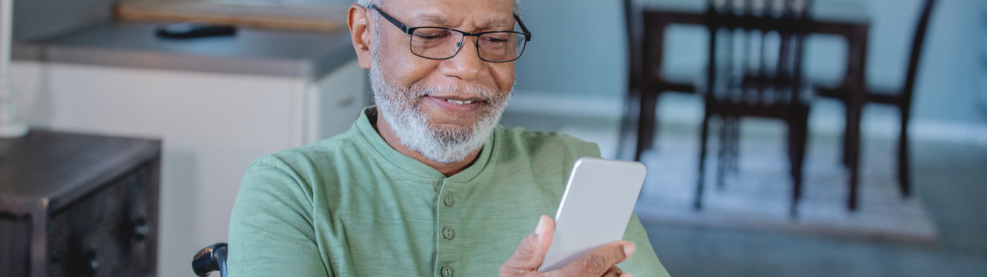 Elderly man in glasses smiles while looking at his smartphone. Soft interior backdrop.