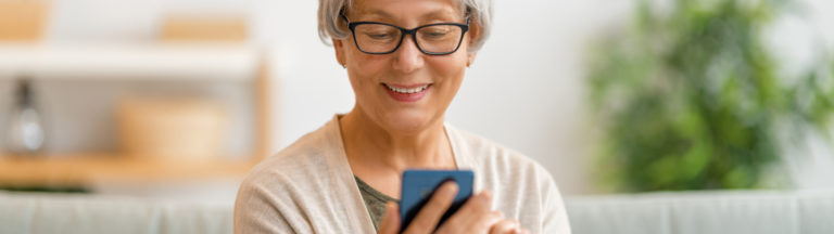 Senior woman with short gray hair, wearing glasses, smiles while using a smartphone.