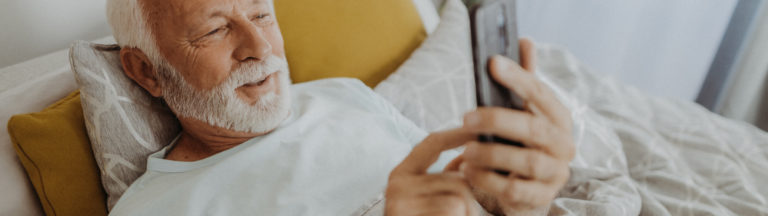 Elderly man in bed, looking at a smartphone with a smile, surrounded by pillows.