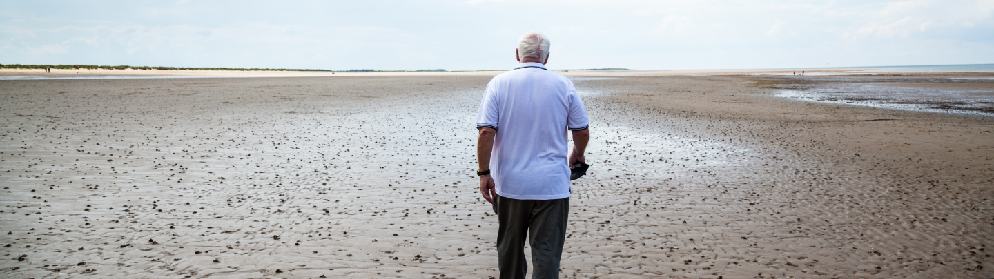 An older man walks alone on a wide, empty beach at low tide under a cloudy sky.