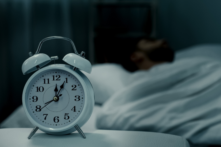 Alarm clock displayed in foreground with a person sleeping under a blanket in the background.