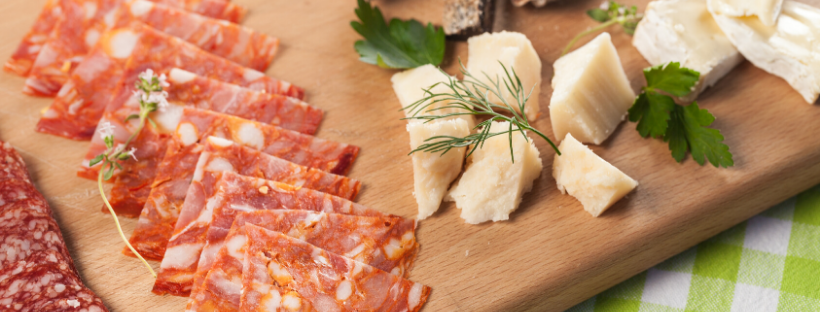 A wooden platter displays sliced salami, cheese cubes, and fresh herbs.