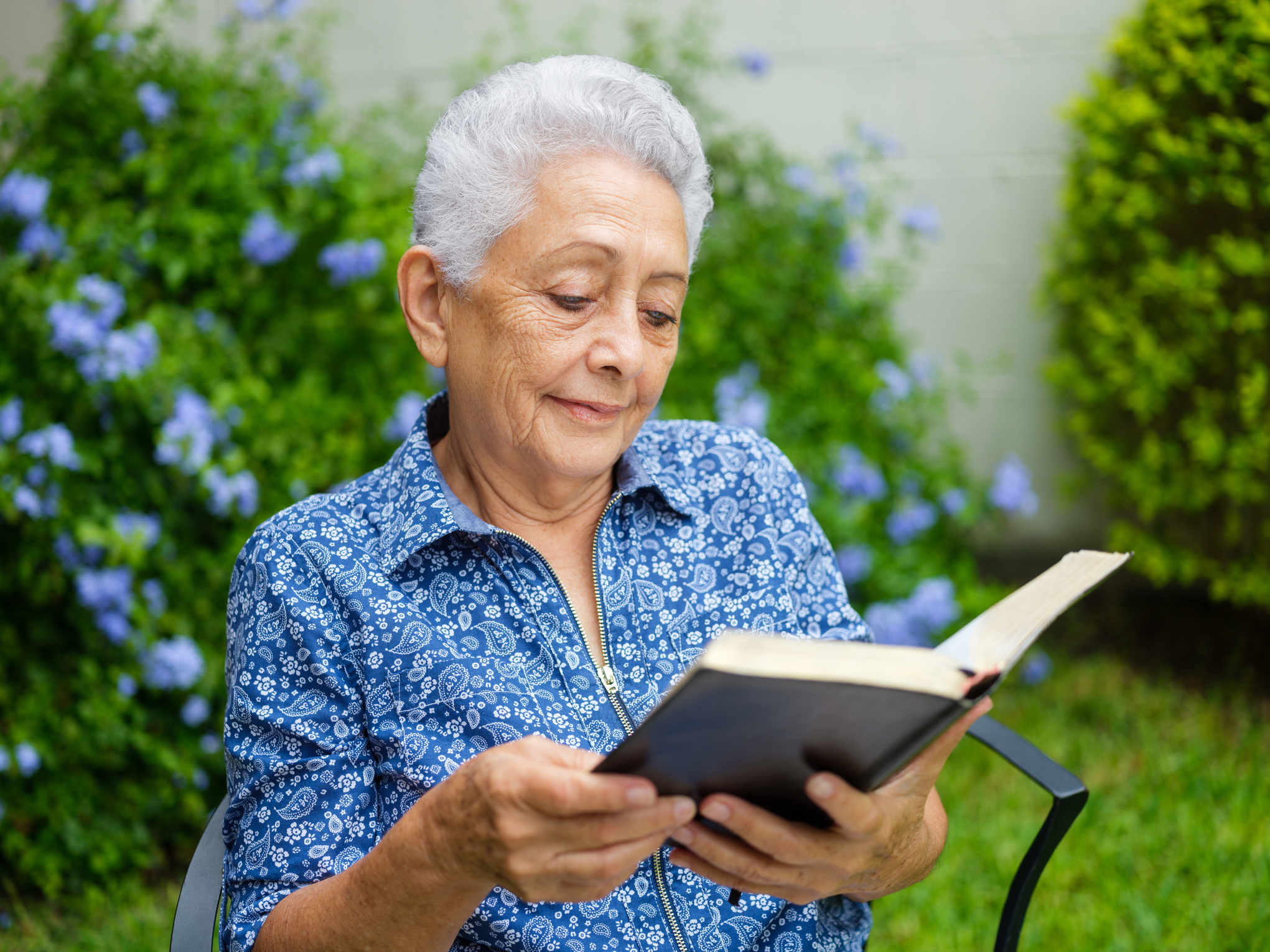 An elderly woman with gray hair reads a book outdoors, surrounded by greenery and flowers.