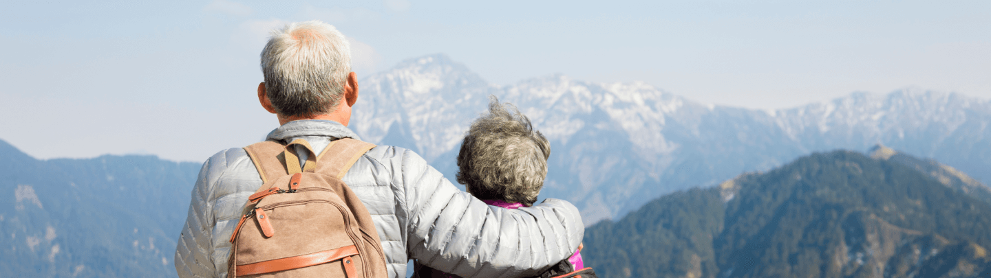 Older couple standing together, overlooking a mountainous landscape with snow-capped peaks.