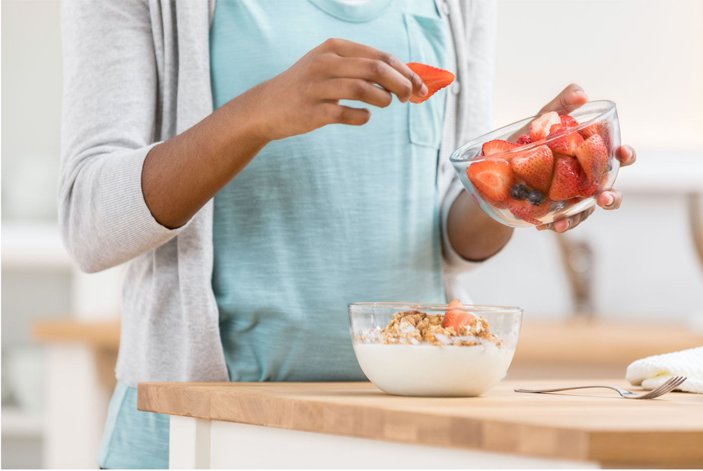 A person adds strawberries to a bowl of yogurt and granola on a wooden countertop.