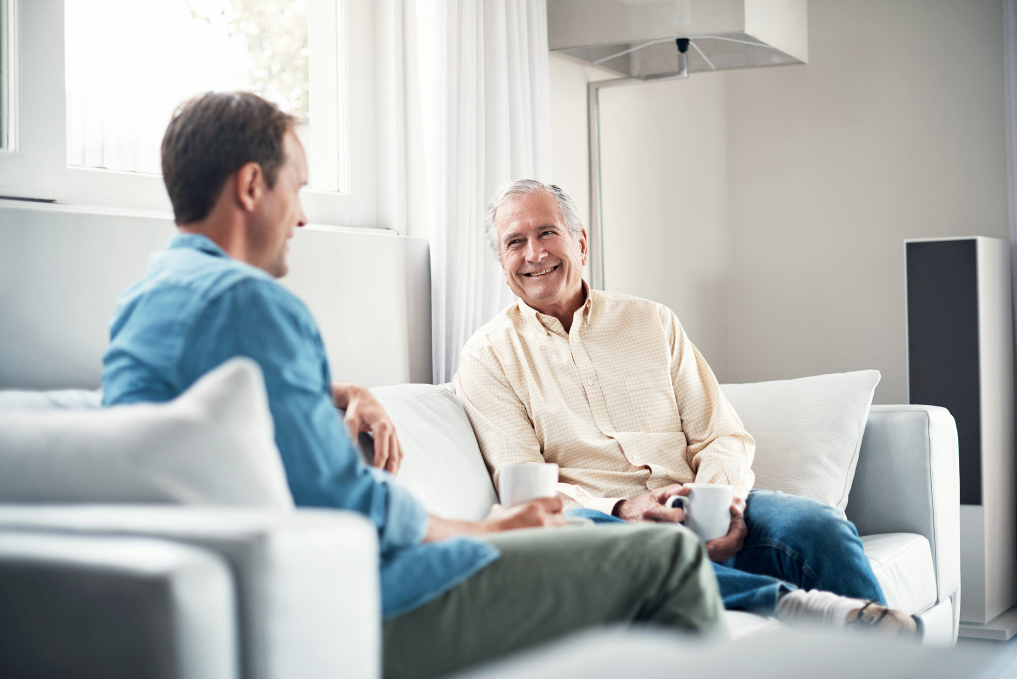 Two men sit on a couch, smiling and having a conversation while holding cups.