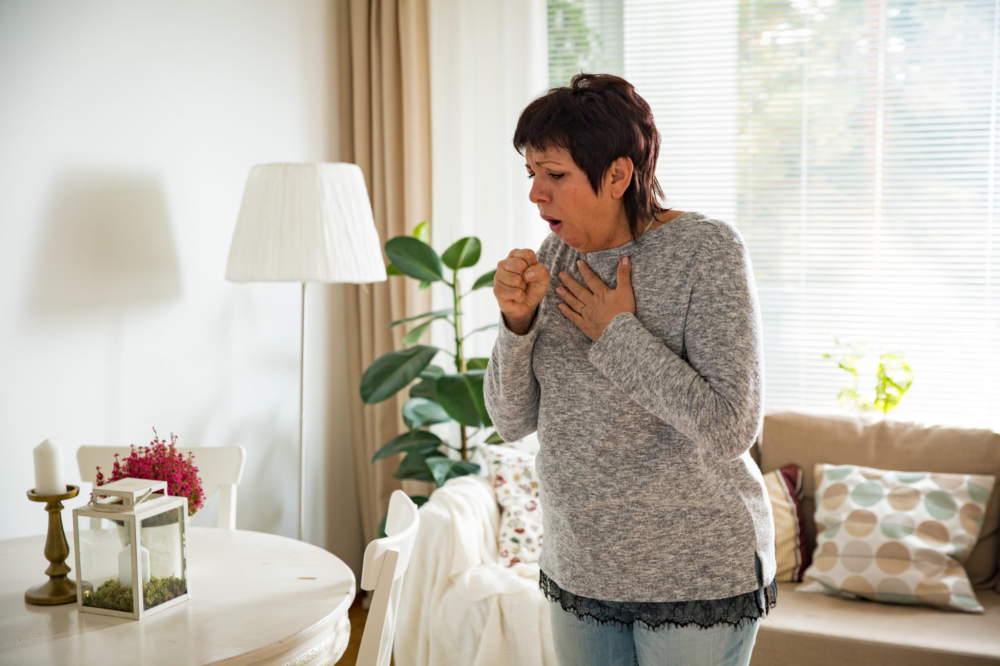 A woman coughs and appears concerned while standing in a cozy living room.