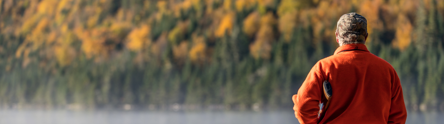 A person in an orange jacket stands by a lake, surrounded by autumn foliage and trees.