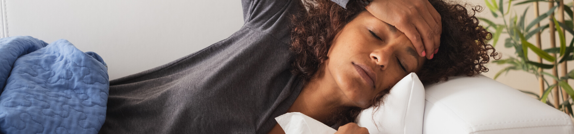 A person with curly hair rests on a couch, holding their forehead in discomfort, possibly feeling