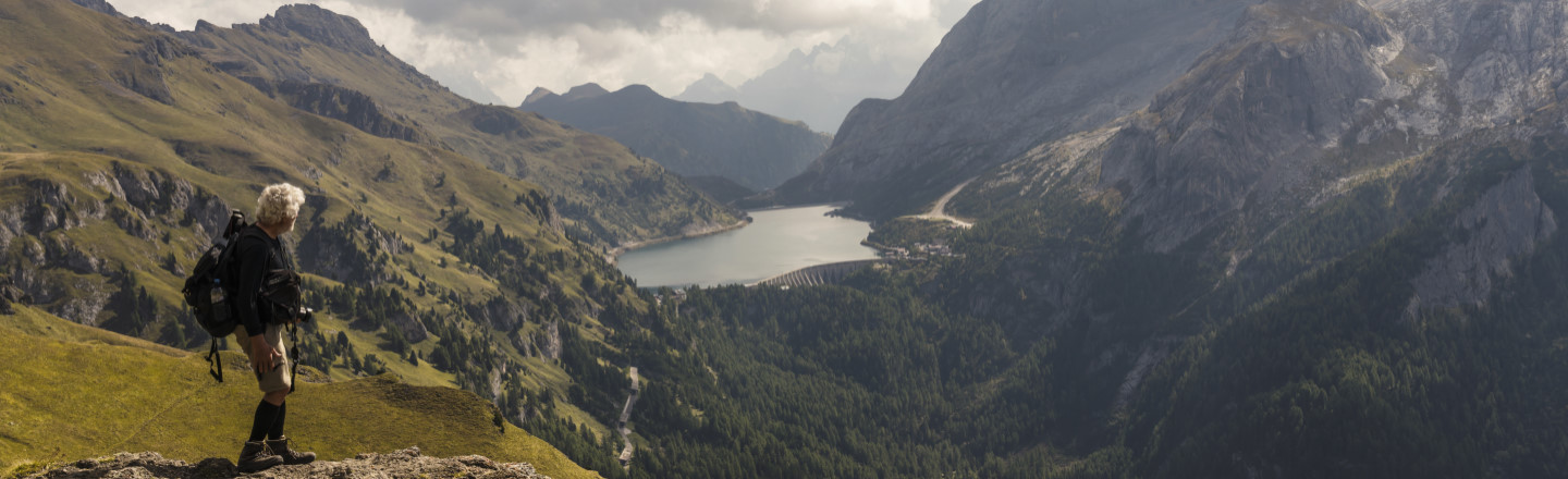 A hiker stands on a rocky ledge overlooking a lush valley and lake, surrounded by mountains.