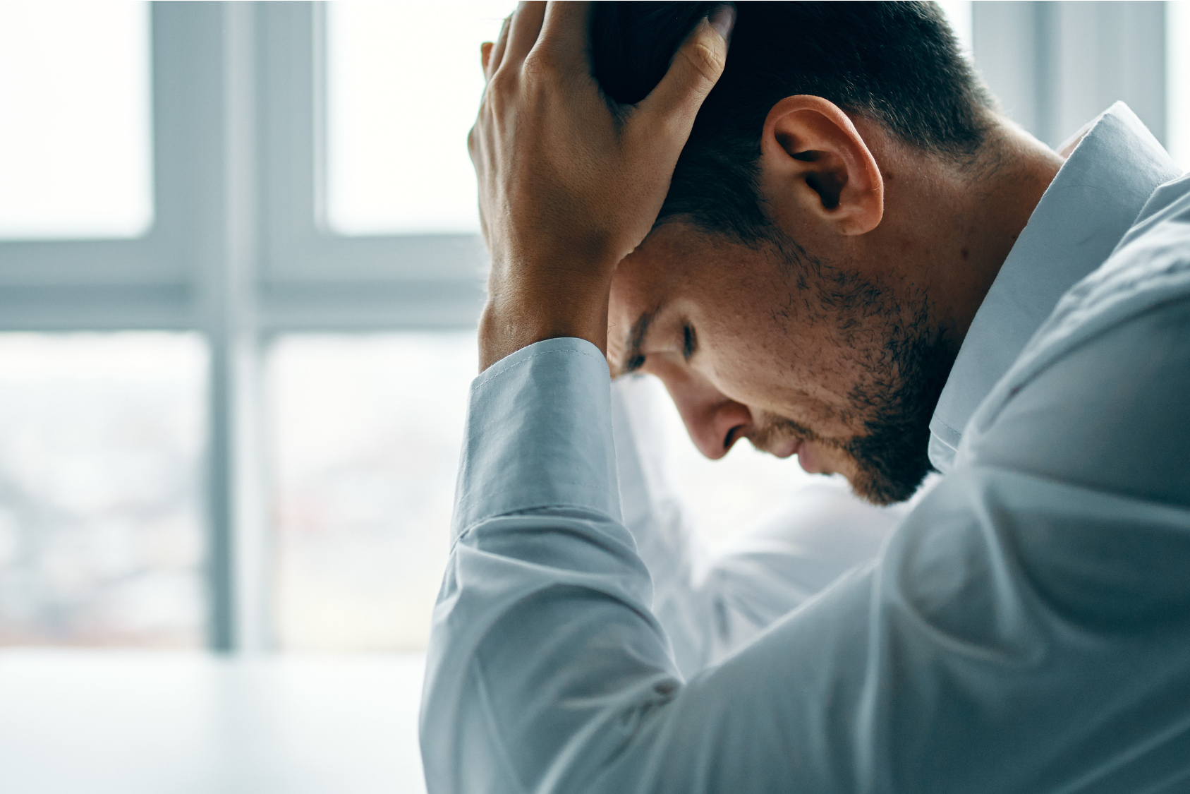 A man in a white shirt sits with his head in his hands, looking stressed or overwhelmed.