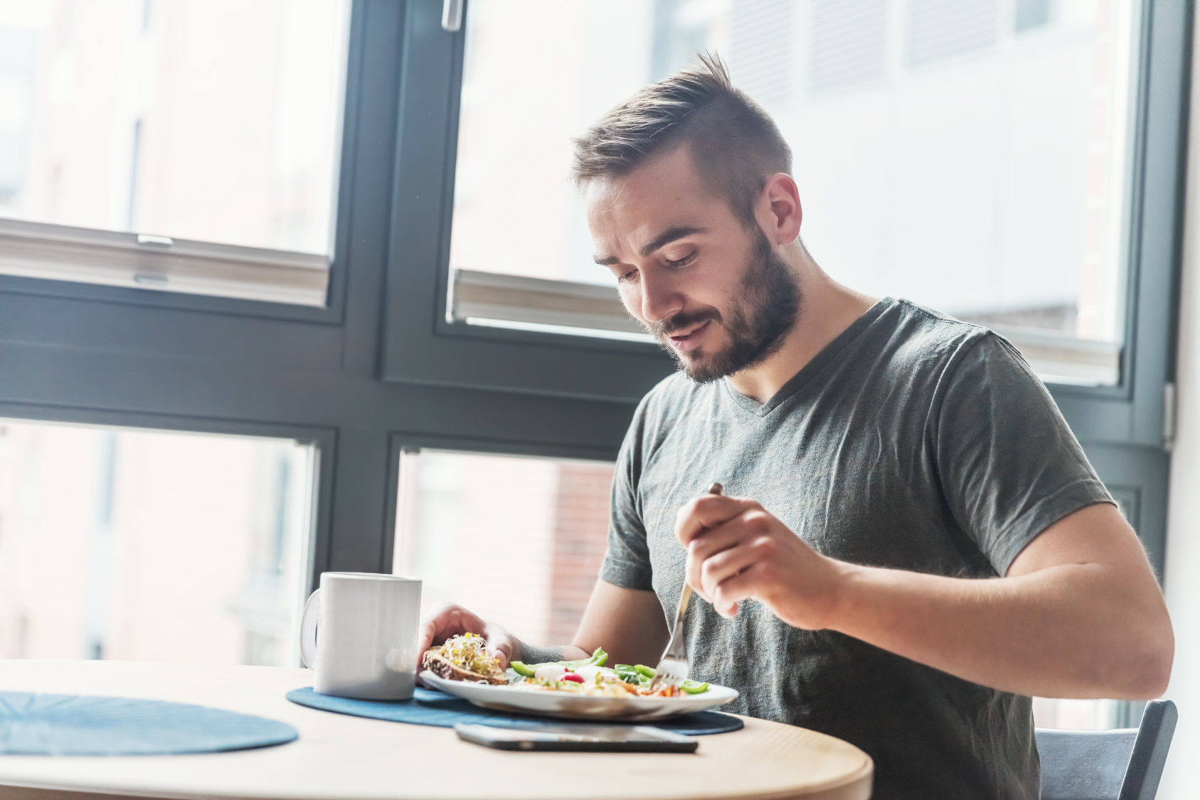 A man enjoys a healthy meal at a table near a window, smiling and using a fork.