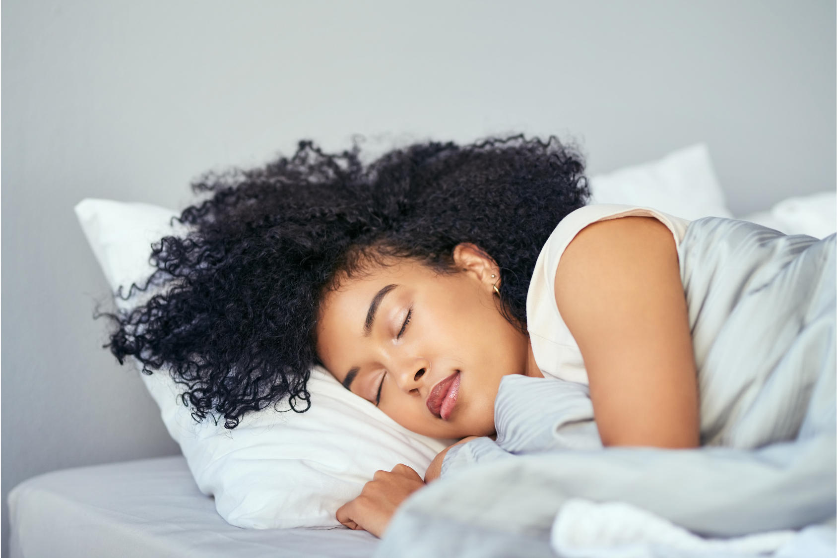 A woman with curly hair sleeps peacefully on a bed with white pillows and sheets.