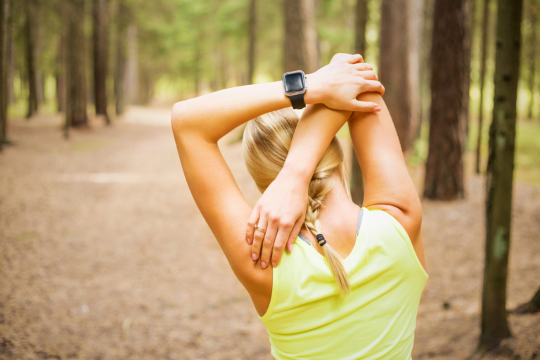 A person stretches their arms in a forest setting, wearing a fitness watch and a yellow tank top.