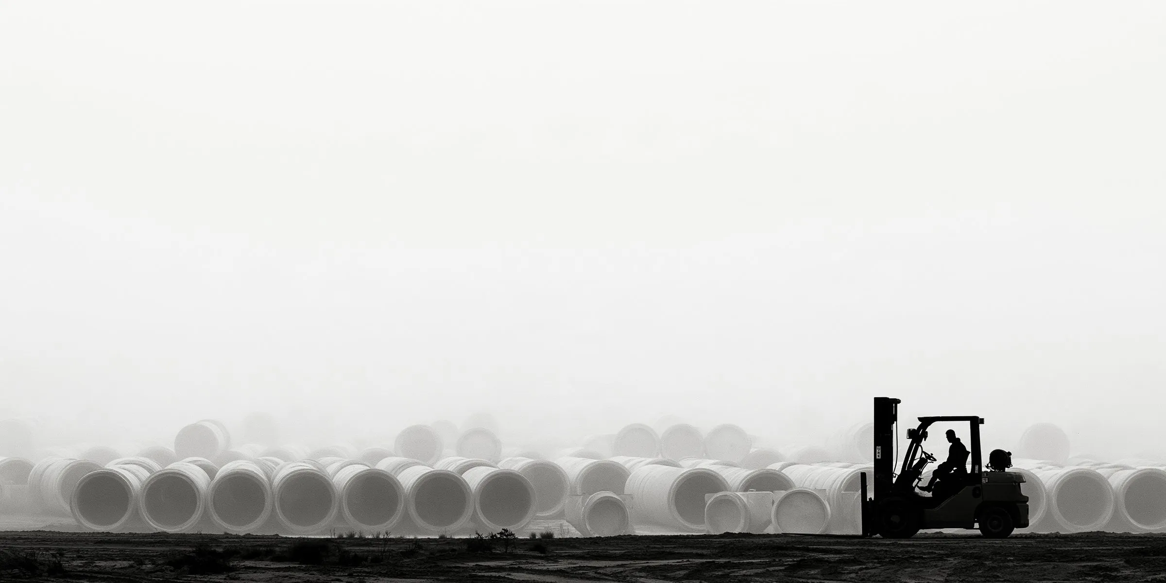 A forklift operator working in a pipe yard under a hazy sky, rows of large white pipe behind