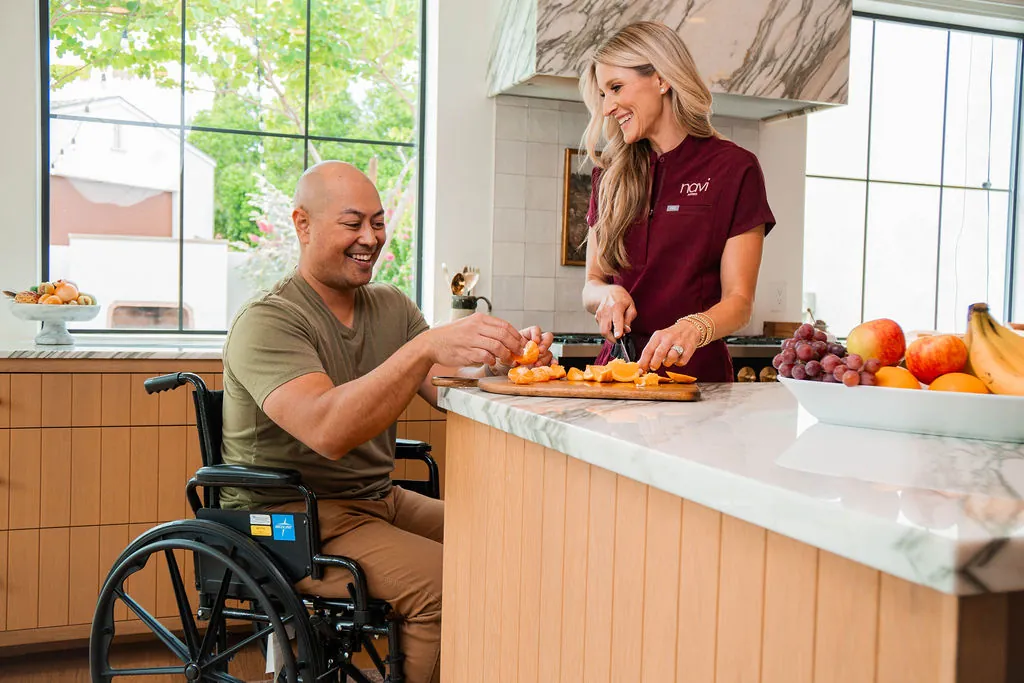A Navi Caregiver slices oranges at a kitchen island with a laughing man in a wheelchair