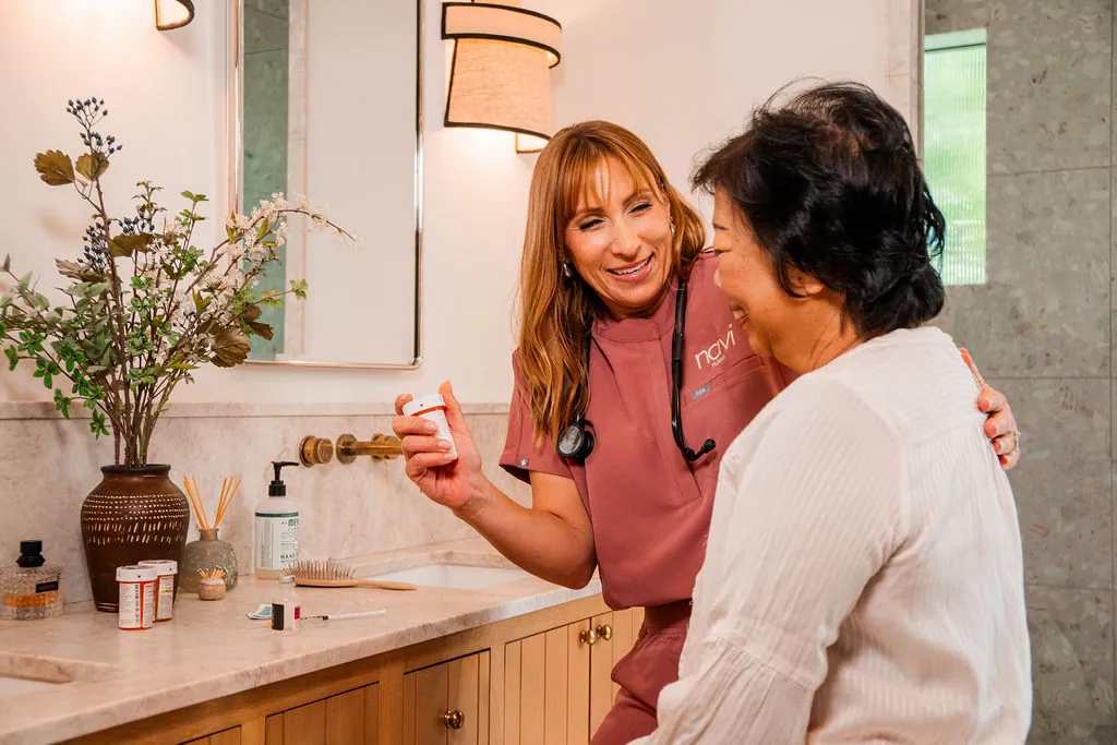 A Navi Nurse with a stethoscope explains a prescription bottle to a smiling woman in her bathroom