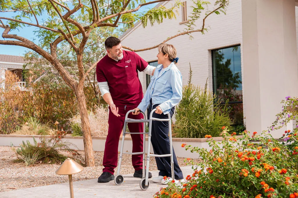 A Navi Caregiver walks beside a woman using a walker along a sunny garden path
