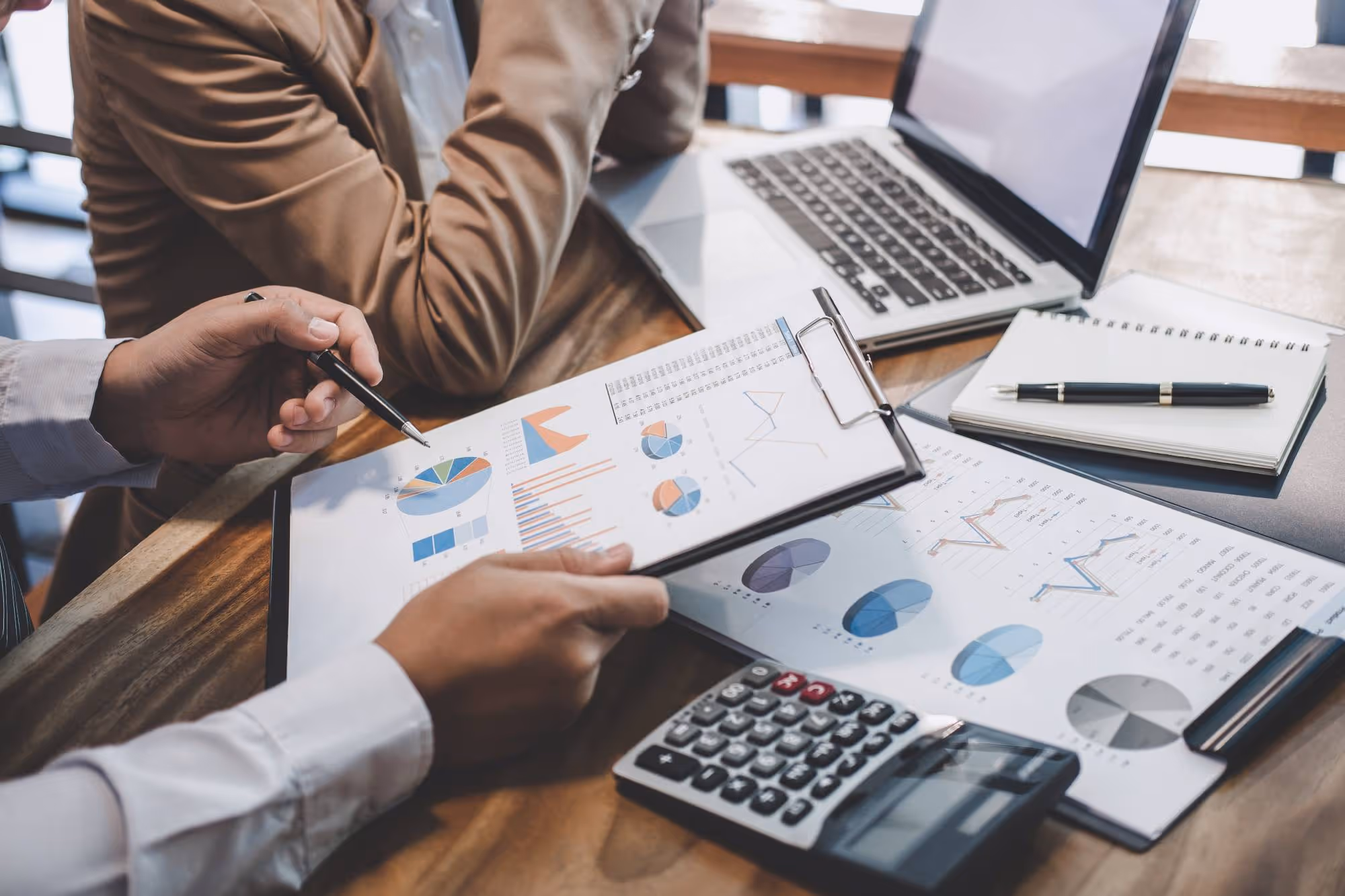 Two professionals reviewing financial charts and graphs on clipboards with a calculator and laptop on a wooden desk.
