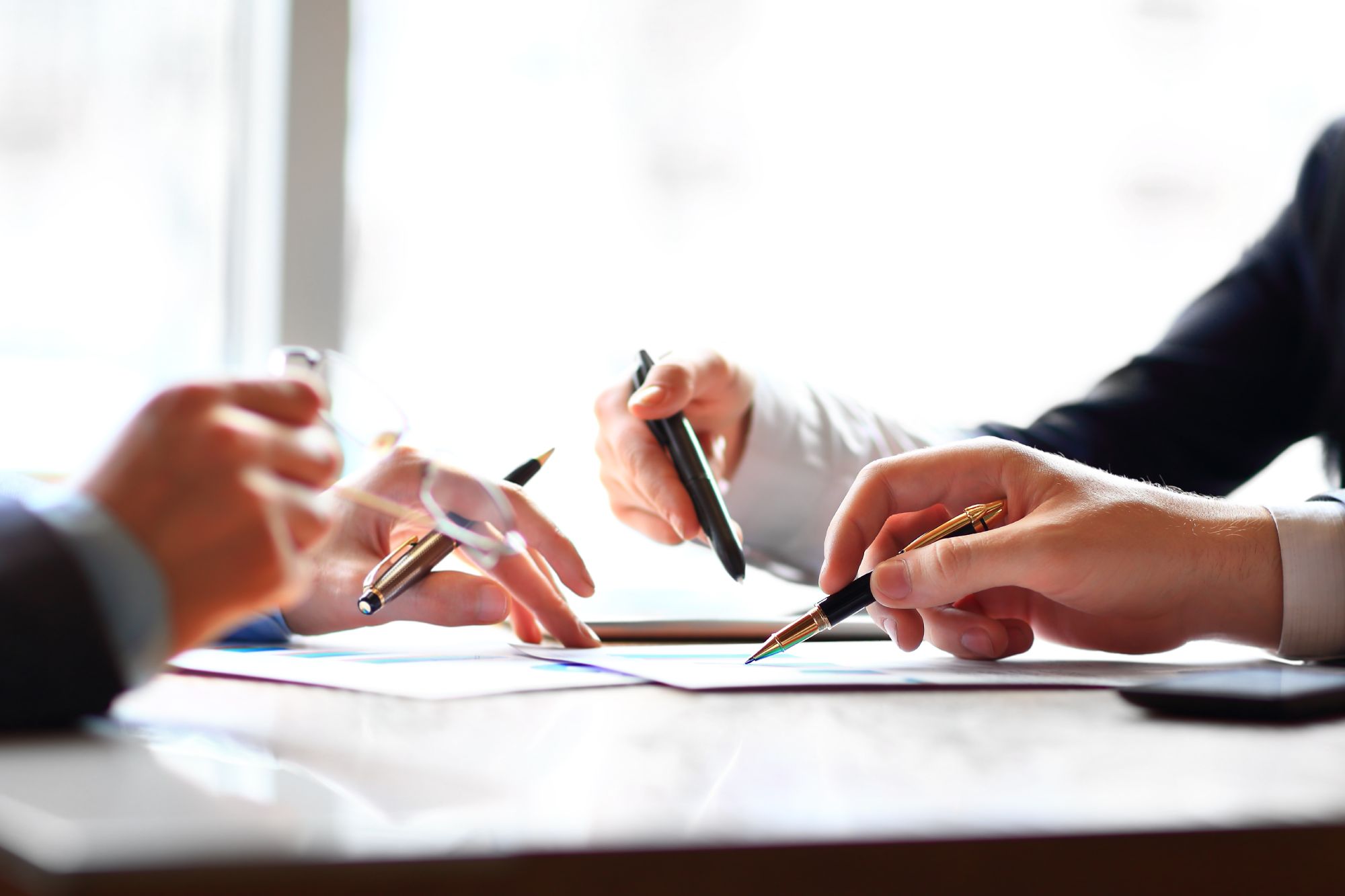 Three hands holding pens and discussing documents on a table in a bright office setting.
