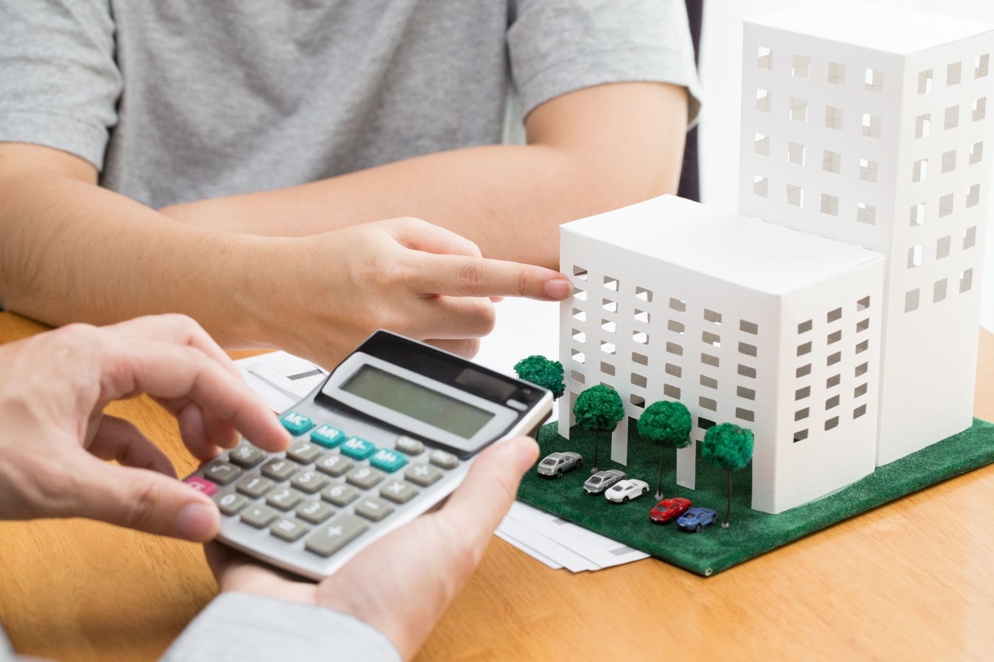 Person pointing at a white architectural model of buildings with miniature trees and cars while another holds a calculator over documents on a wooden table.
