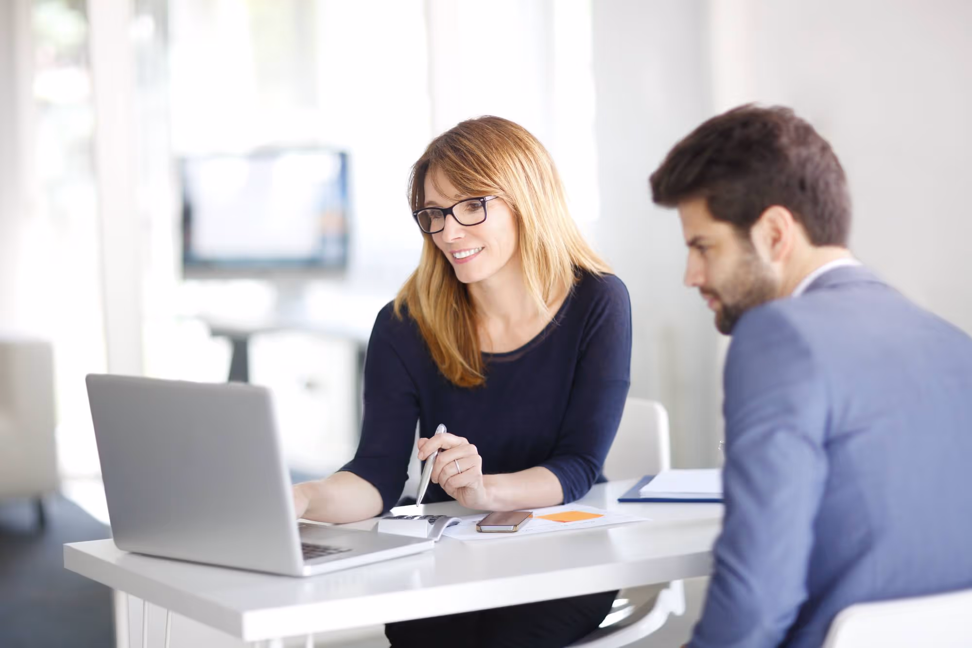 Smiling woman with glasses explaining something on a laptop to a man in a blue suit at a white desk.